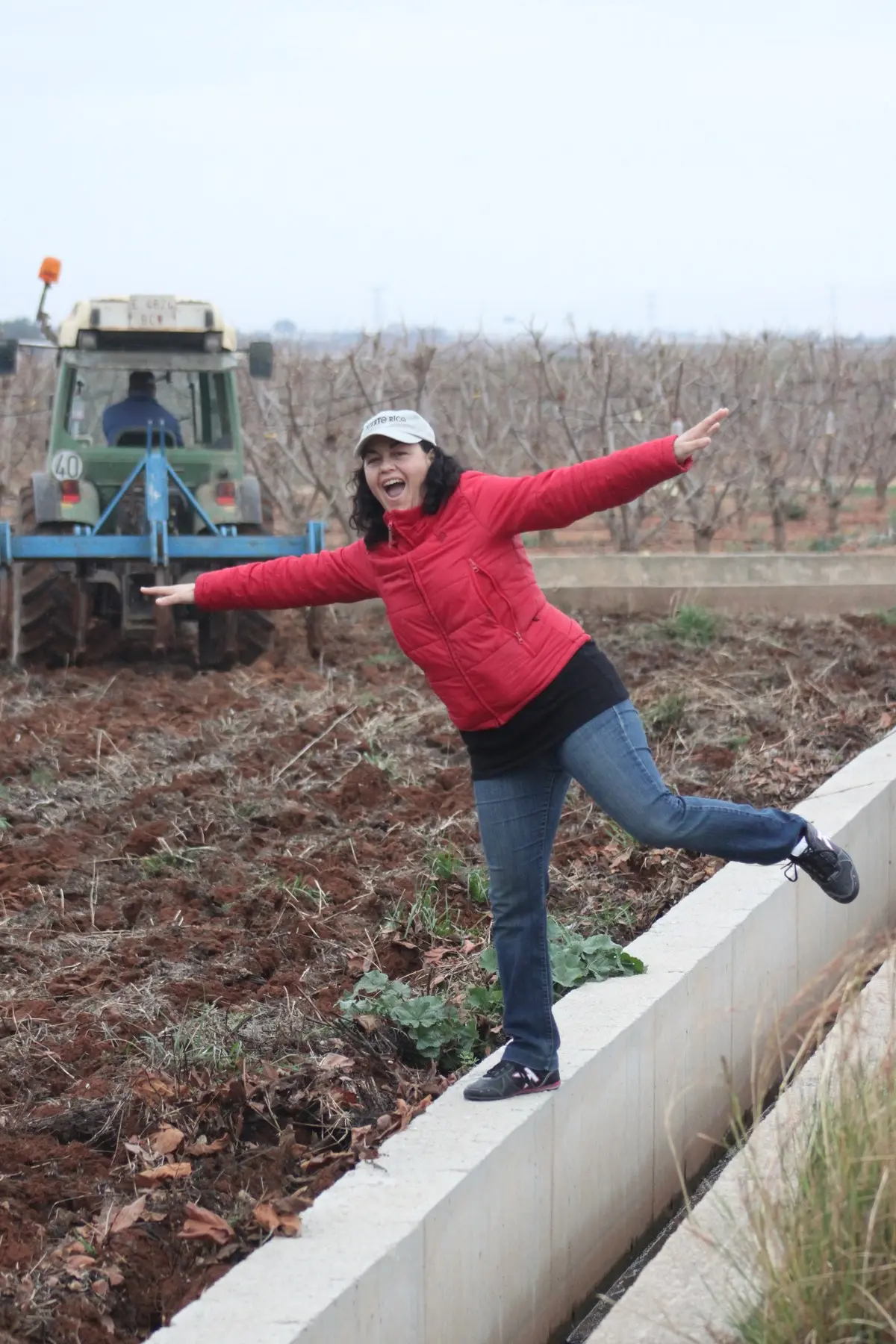 Isabel sobre el borde de una acequia mientras un tractor pasa los pinchos en el campo donde se cultivarían los primeros chiles mexicanos en España